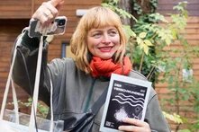 Eine weiße, blonde Frau um die 40 gestikuliert ausschweifend während sie etwas einem Publikum ausserhalb der Bildes zu erklären scheint. Sie sieht freundlich und etwas verschmitzt aus. Ihre Haare sind zu einem Bob mit geradem Pony geschnitten. Ihre Augen sind grünblau. Sie trägt roten Lippenstift, eine Bomberjacke und einen orangen Schal.