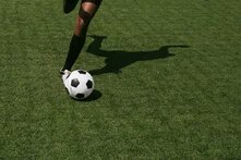 Ein Bein spielt einen Fussball. Auf das Fussballfeld fällt der Schatten des*r Spielenden.