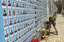 Foto: Eine Gedenkwand mit zahlreichen Porträtfotos verstorbener Soldat*innen. Davor stehen Blumen und Kränze. Ein Gehweg führt entlang der Wand.