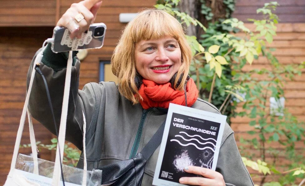 Eine weiße, blonde Frau um die 40 gestikuliert ausschweifend während sie etwas einem Publikum ausserhalb der Bildes zu erklären scheint. Sie sieht freundlich und etwas verschmitzt aus. Ihre Haare sind zu einem Bob mit geradem Pony geschnitten. Ihre Augen sind grünblau. Sie trägt roten Lippenstift, eine Bomberjacke und einen orangen Schal.