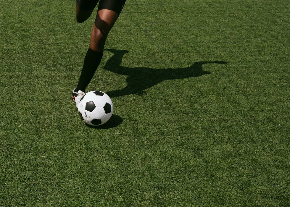 Ein Bein spielt einen Fussball. Auf das Fussballfeld fällt der Schatten des*r Spielenden.