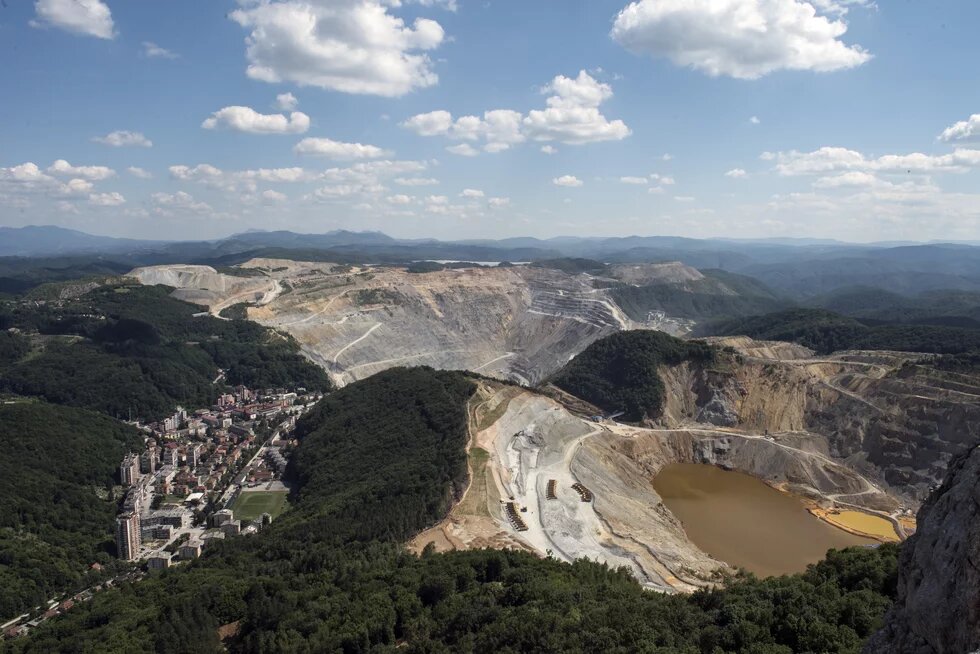 Photo: Mining area near Majdanpek, Serbia, in a hilly landscape. In the foreground is a town with many buildings. On the right, a large open-pit mine with a water basin.